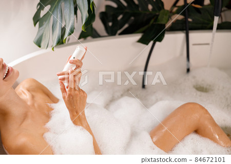 Relaxed young woman smiling while taking bubble bath and holding body care product, taking care of herself at luxury spa resort 84672101
