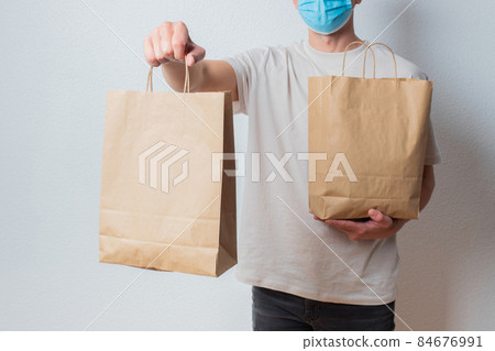 Young delivery man in protective face mask holding paper bags in hands. Man courier with shopping bag. Safe online delivery from supermarket to home. COVID-19 coronavirus protection Young delivery man in protective face mask holding paper bags in hands. Man courier with shopping bag. Safe online delivery from supermarket to home. COVID-19 coronavirus protection 84676991