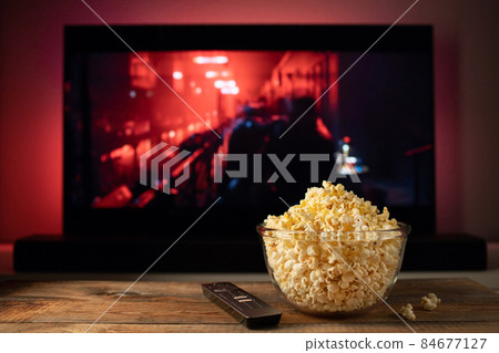 A glass bowl of popcorn and remote control in the background the TV works. Evening cozy watching a movie or TV series at home 84677127