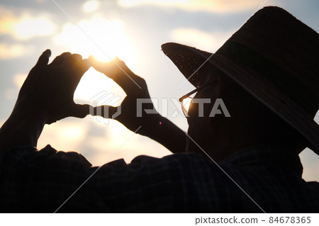 Silhouette of Senior farmer in a hat looking into the distance at sunset and making a heart shape on a golden sky background. 84678365