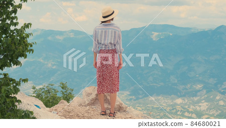 Happy traveler beautiful mountains view. Woman in hat, red dress celebrates climb, against mountain valley background. Happy traveler enjoys mountain view Happy traveler beautiful mountains view. Woman in hat, red dress celebrates climb, against mountain valley background. Happy traveler enjoys mountain view 84680021