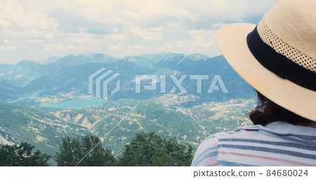 Close-up, woman traveler looking at mountains view from top. Happy woman traveler portrait in straw hat watching at scenery summer mountain landscape. Travel, trekking concept. Albania, Dajti 84680024
