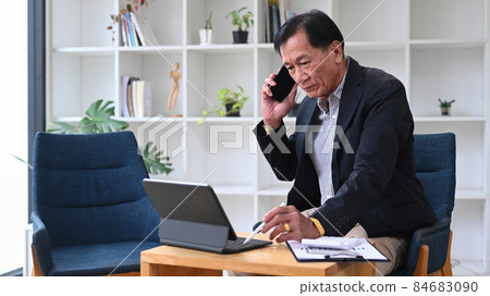 Mature businessman talking on mobile phone and working with computer tablet in his office room. Mature businessman talking on mobile phone and working with computer tablet in his office room. 84683090