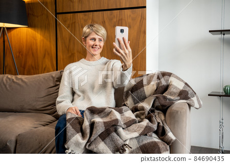 A middle-aged woman is sitting on the sofa in the living room, talking via video on a modern cell phone gadget, an older woman greeting, talking on the Internet using a smartphone 84690435