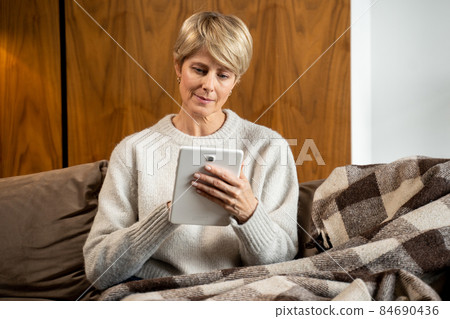 Portrait of a middle-aged woman sitting on a cozy sofa and using a tablet computer at home. A woman shopping in a popular online store 84690436