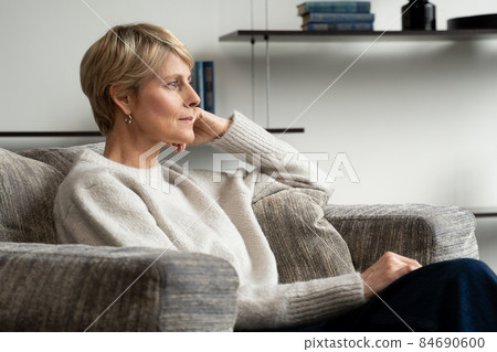 A middle-aged woman is sitting alone in an armchair and looking at the view from the window. 84690600
