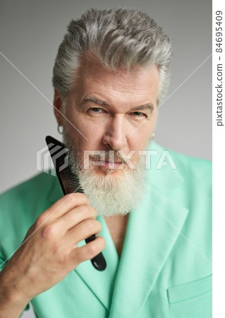 Portrait of attractive gray haired mature man looking at camera, brushing his beard with comb, posing over white background Portrait of attractive gray haired mature man looking at camera, brushing his beard with comb, posing over white background 84695409