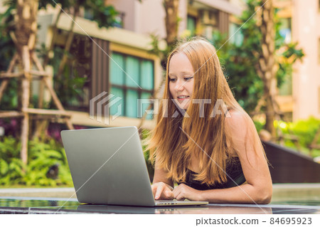 Young female freelancer sitting near the pool with her laptop in the hotel browsing in her smartphone. Busy at holidays. Distant work concept. Copy space for your text Young female freelancer sitting near the pool with her laptop in the hotel browsing in her smartphone. Busy at holidays. Distant work concept. Copy space for your text 84695923