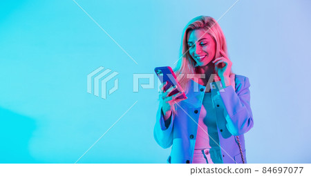 Happy fashionable young girl with a beauty smile in fashion elegant business clothes with a jacket holding a smart phone and typing a message on a multicolored neon pink light in the studio 84697077