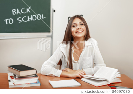 Happy beautiful young girl teacher with a smile sits at a desk with textbooks in the classroom near the blackboard with chalk inscription Back to school, start of studies 84697080