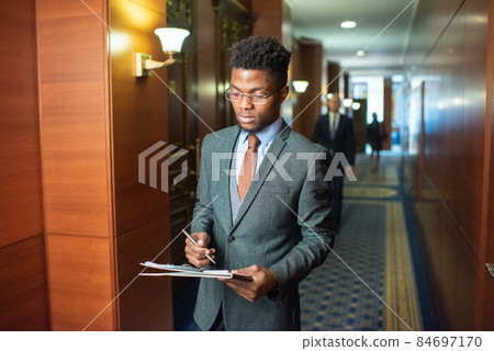 Young African businessman with clipboard standing in corridor 84697170