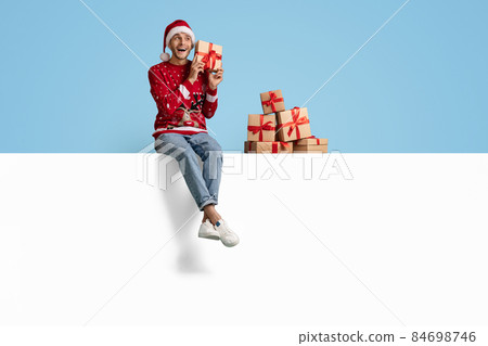 Holiday Offer. Excited Man With Gift Boxes Sitting On White Advertisement Board 84698746