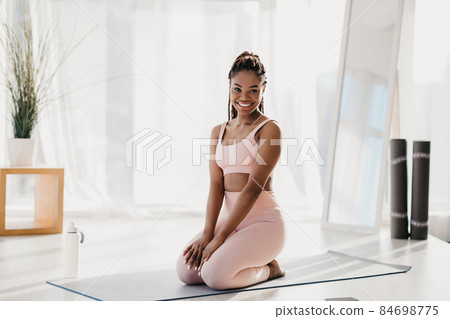Smiling young black woman exercising at home, taking break, resting on yoga mat indoors, full length 84698775