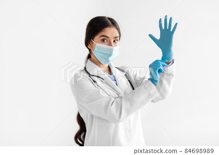 Serious millennial indian female doctor in coat and protective mask putting on hand rubber glove 84698989