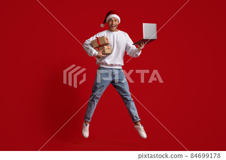 Man In Santa Hat With Laptop And Gifts Jumping Over Red Background 84699178