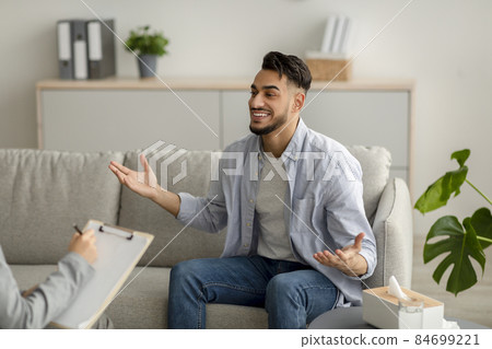 Young happy arab guy talking to his psychologist, receiving professional help, sitting on sofa at mental health clinic 84699221