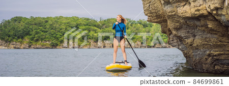 BANNER, LONG FORMAT Young women Having Fun Stand Up Paddling in the sea. SUP. Red hair girl Training on Paddle Board near the rocks 84699861