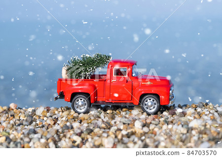 Red retro small car carrying christmas tree on the sea beach shore in snowfall. Winter holiday concept Red retro small car carrying christmas tree on the sea beach shore in snowfall. Winter holiday concept 84703570