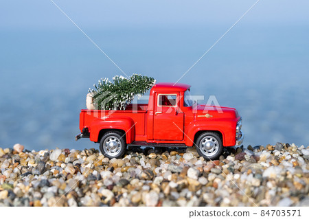 Red retro small car carrying christmas tree on the sea beach shore. Winter holiday concept 84703571