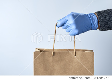 A hand in a blue rubber glove holds out a paper bag on a gray background. Safe shopping concept. Photo with copy space. A hand in a blue rubber glove holds out a paper bag on a gray background. Safe shopping concept. Photo with copy space. 84704238