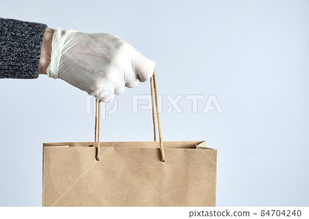 Hand in white rubber gloves holds a paper bag. Safe shopping concept. Food delivery during the quarantine of the coronavirus pandemic. Hand in white rubber gloves holds a paper bag. Safe shopping concept. Food delivery during the quarantine of the coronavirus pandemic. 84704240