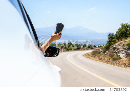 Woman driving a car holding paper cup of coffee from the open window. Trip on the serpentine road in the mountains. Summer vacation Woman driving a car holding paper cup of coffee from the open window. Trip on the serpentine road in the mountains. Summer vacation 84705892