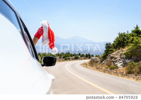 Woman in christmas gloves driving a car holding santa claus hat from the open window. Trip on the serpentine road in the mountains. 84705928