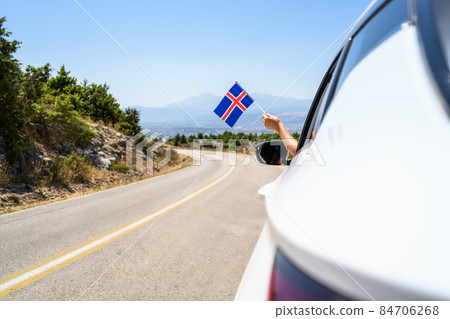 Woman holding Iceland flag from the open car window driving along the serpentine road in the mountains. Concept 84706268