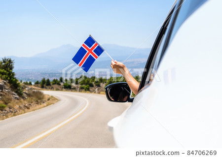Woman holding Iceland flag from the open car window driving along the serpentine road in the mountains. Concept 84706269