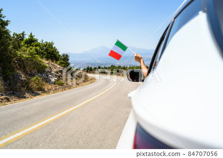 Woman holding  Italy flag from the open car window driving along the serpentine road in the mountains. Concept 84707056