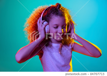 Portrait of little girl, kid listening to music in headphones isolated on blue studio background in neon light, filter. 84707713