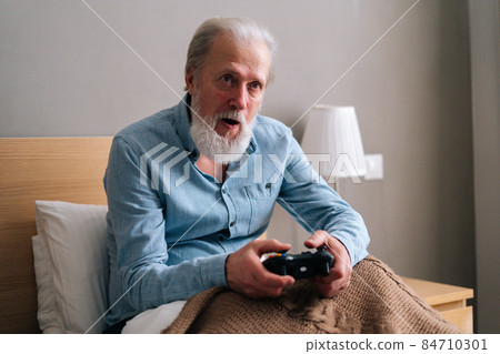 Front view of focused gray-haired senior male playing online video game using gamepad at home, having fun alone. 84710301