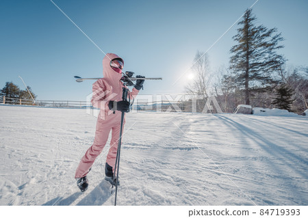 Winter Ski. Skiing portrait of woman alpine skier holdings skis wearing helmet, cool ski goggles, hardshell winter jacket and ski gloves on cold day in front of snow covered trees on ski trail slope 84719393