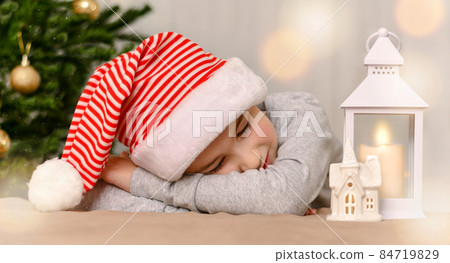 A child who fell asleep under the Christmas tree in a cap. With a happy smile and closed eyes, he lay down on hands. 84719829