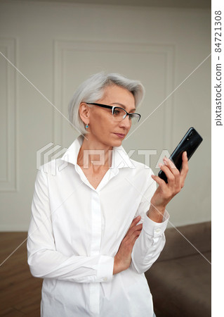 Mature confident female in glasses looking at screen of smartphone Mature confident female in glasses looking at screen of smartphone 84721308