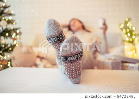 Woman sitting on couch near christmas tree. Female legs, feet in winter woolen socks putting on the table and relaxing after packing handmade gift boxes. Presents for family. Woman sitting on couch near christmas tree. Female legs, feet in winter woolen socks putting on the table and relaxing after packing handmade gift boxes. Presents for family. 84722483
