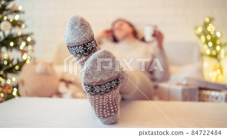 Woman sitting on couch near christmas tree. Female legs, feet in winter woolen socks putting on the table and relaxing after packing handmade gift boxes. Presents for family. Woman sitting on couch near christmas tree. Female legs, feet in winter woolen socks putting on the table and relaxing after packing handmade gift boxes. Presents for family. 84722484