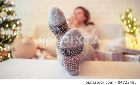 Woman sitting on couch near christmas tree. Female legs, feet in winter woolen socks putting on the table and relaxing after packing handmade gift boxes. Presents for family. Woman sitting on couch near christmas tree. Female legs, feet in winter woolen socks putting on the table and relaxing after packing handmade gift boxes. Presents for family. 84722485