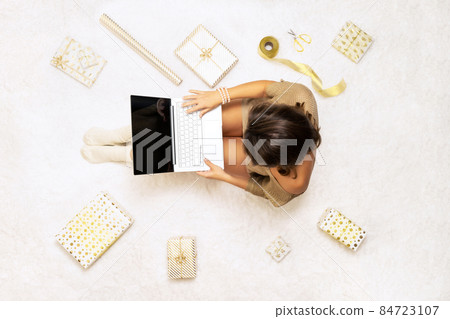 Christmas. Online shopping. Woman in sweater using laptop for making order, buying, sitting on the white carpet among the many wrapped gifts in white gold wrapping paper. Top view. Concept 84723107
