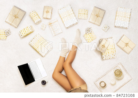 Christmas sale, discount. Online shopping. Woman using laptop for making order, buying gifts, sitting on the snow-white carpet among many wrapped boxes. Top view. Concept 84723108