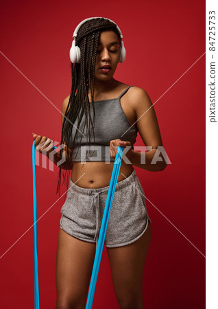 Young athletic African woman with Afro pigtails wearing wireless headphones and gray top and shorts, exercising with fitness elastic strap against red colored background with copy space 84725733