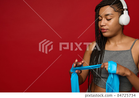 Close-up portrait of an African sportswoman wearing wireless headphones wrapping a blue elastic strap on her hand while standing on a red background with a copy of space 84725734