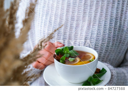 Female hands on sweater holding tea with lemon, cinnamon sticks and mint leaves. Hot drink. Healthy melissa tea natural organic aromatic drink in cup. Autumn winter warming drink. Cozy home 84727388