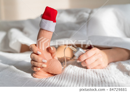 A woman lies in bed and holds a dildo with a control panel in a santa claus hat A woman lies in bed and holds a dildo with a control panel in a santa claus hat 84729881