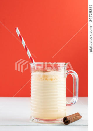 Traditional Christmas hot drink eggnog made of milk, eggs and cinnamon in glass mug with drinking straw on wooden table on red backdrop. Holiday beverage minimal image. Copy space. Vertical shot. Traditional Christmas hot drink eggnog made of milk, eggs and cinnamon in glass mug with drinking straw on wooden table on red backdrop. Holiday beverage minimal image. Copy space. Vertical shot. 84729882
