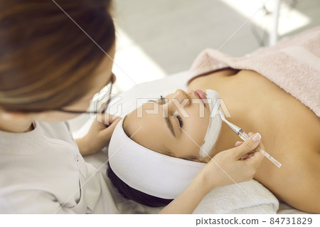 Professional cosmetologist at spa center applying anti acne kaolin clay mask on girl's face. Calm woman lying on bed in beauty parlor getting cosmetic facial treatment for fresh, beautiful, clear skin Professional cosmetologist at spa center applying anti acne kaolin clay mask on girl's face. Calm woman lying on bed in beauty parlor getting cosmetic facial treatment for fresh, beautiful, clear skin 84731829