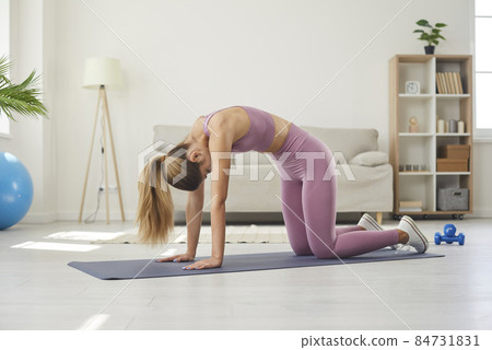 Beautiful fit young woman having fitness and yoga workout at home. Caucasian girl practicing yoga on exercise mat and doing Cat and Cow pose to stretch and loosen her back and warm up her muscles 84731831