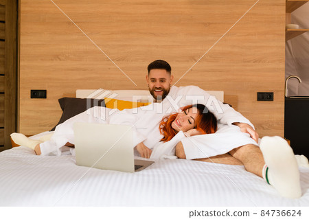 Couple in bathrobes is lying on bed with a laptop. 84736624