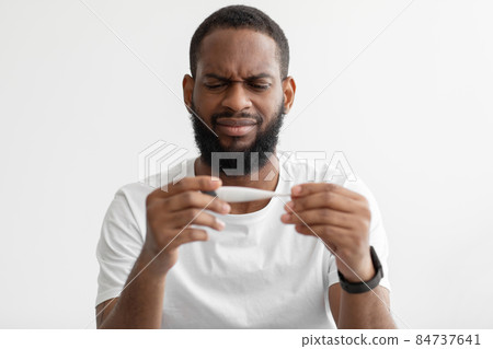 Portrait of unhappy millennial black bearded man look at thermometer with high temperature on white wall background Portrait of unhappy millennial black bearded man look at thermometer with high temperature on white wall background 84737641