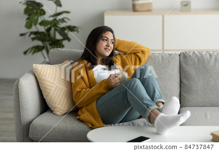 Peaceful young arab woman relaxing on sofa with cup of coffee, looking aside and dreaming, free space 84737847